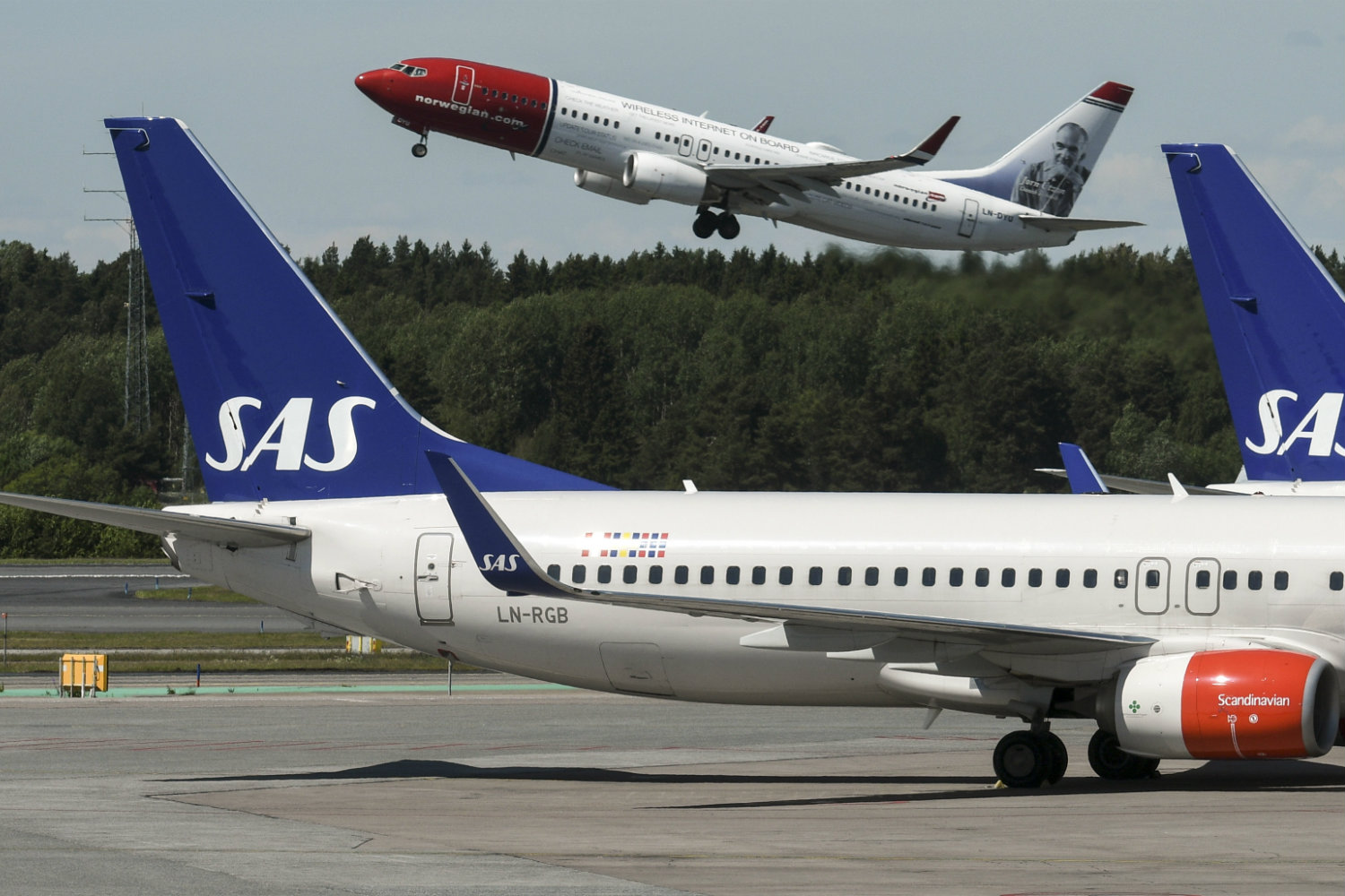 Due aerei della compagnia Sas parcheggiati in primo piano; dietro un velivolo di Norwegian Air che decolla